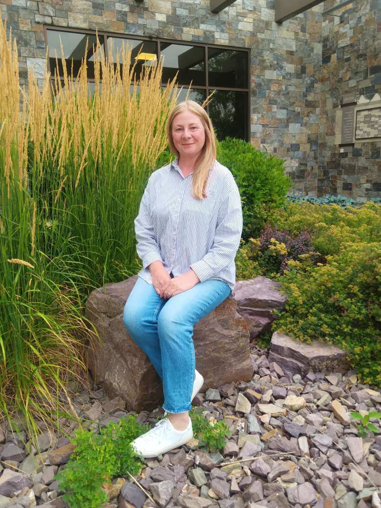 Smiling Nicole Vranizan, Board of Directors, sitting outside of the Hospital on a rock.